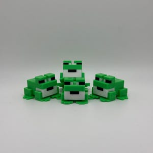 Pode incluir: Cinco figuras de sapos verdes e brancos em blocos estão dispostas sobre uma superfície branca. Os sapos têm olhos pretos e estão num estilo divertido e de desenho animado. As figuras são feitas de um material sólido.