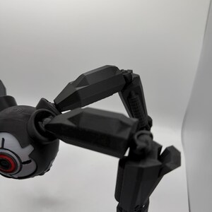 May include: A black, articulated robotic spider with a round head and multiple jointed legs. The head features a gray and red eye-like design. The legs are segmented and angular, suggesting a mechanical or futuristic theme.