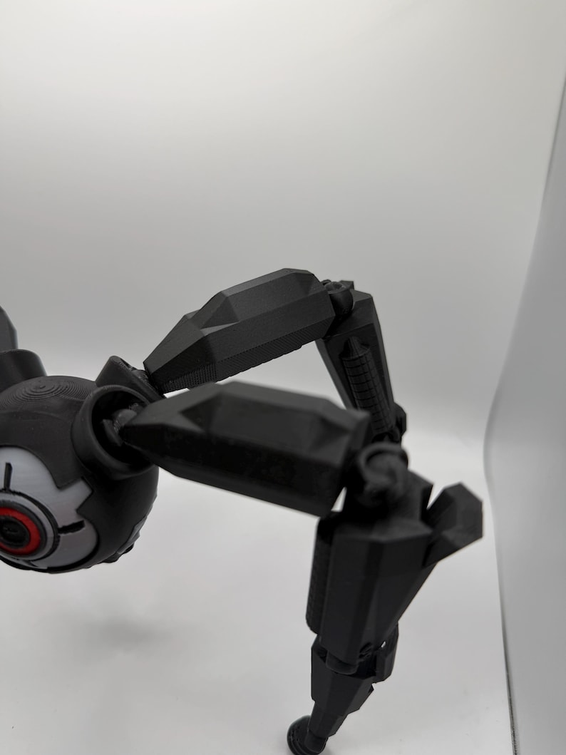 May include: A black, articulated robotic spider with a round head and multiple jointed legs. The head features a gray and red eye-like design. The legs are segmented and angular, suggesting a mechanical or futuristic theme.