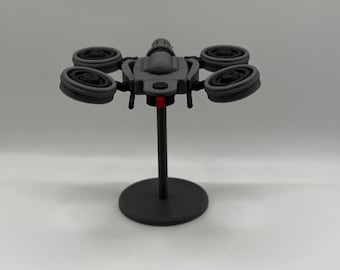 May include: A gray drone model with four propellers, mounted on a black stand. The drone has a central body with a red accent and is designed with detailed features. The stand has a circular base.