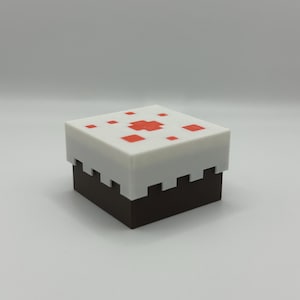 May include: A square, two-tiered cake-shaped box. The top tier is white with red square accents, and the bottom tier is dark brown. The top has a pixelated design, reminiscent of a video game.