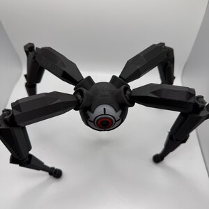 May include: A black, geometric, spider-like robot with eight articulated legs. The central body features a circular design with a red and white eye-like detail. The robot is set against a plain white background.