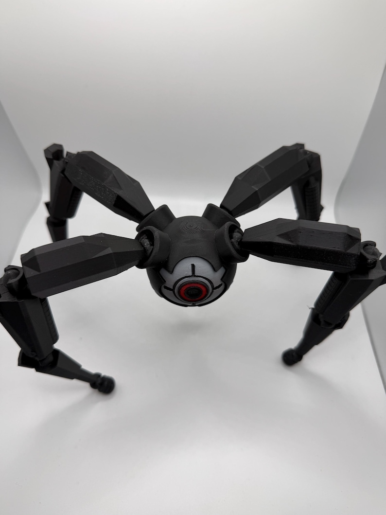 May include: A black, geometric, spider-like robot with eight articulated legs. The central body features a circular design with a red and white eye-like detail. The robot is set against a plain white background.