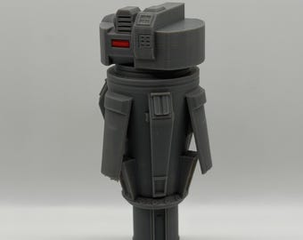 May include: A gray, 3D-printed model of a cylindrical robot with a rectangular head. The head has a red accent and several small rectangular details. The body has several rectangular protrusions. The model is on a white background.