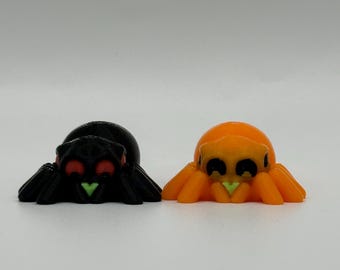 May include: Two small, 3D-printed spider figurines. One is black with red eyes and a green marking, the other is orange with black eyes and a green marking. The spiders have eight legs and are displayed on a white background.
