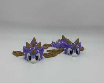 May include: Two purple and white dragon figurines with brown waffle cone wings. The dragons have large eyes and a white face.