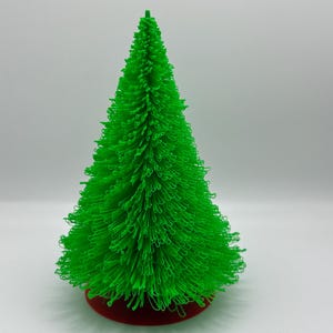 Könnte beinhalten: Ein kleiner, leuchtend grüner Weihnachtsbaum mit einer strukturierten, borstigen Optik. Der Baum steht auf einem runden, roten Sockel. Der Baum hat eine leuchtend grüne Farbe und steht vor einem weißen Hintergrund.