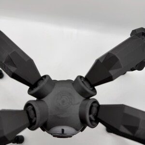 May include: A black, geometric, multi-armed object with a central hub and six angled arms. The object appears to be 3D-printed, with a matte finish and visible layer lines. The arms are symmetrical and pointed.