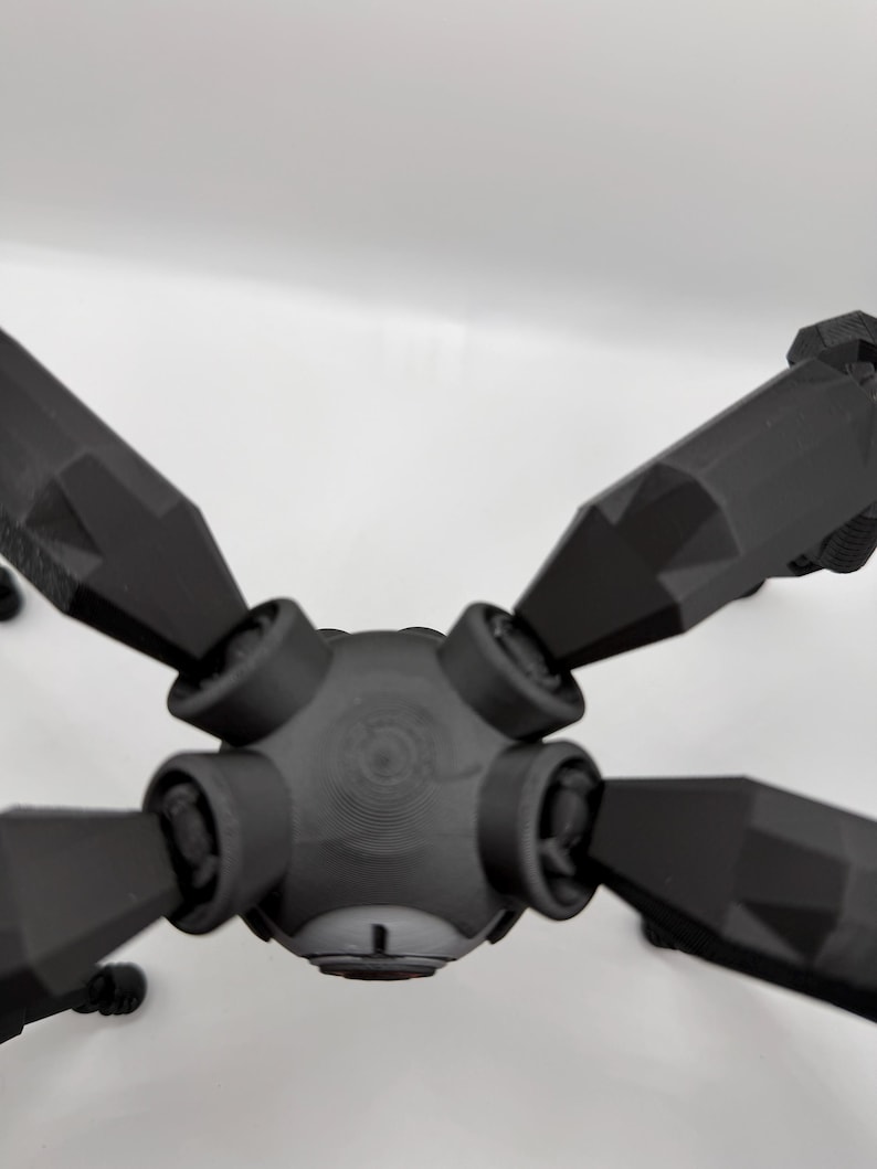 May include: A black, geometric, multi-armed object with a central hub and six angled arms. The object appears to be 3D-printed, with a matte finish and visible layer lines. The arms are symmetrical and pointed.