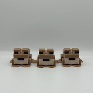 May include: Three pixelated frog figurines in tan, brown, and black. The frogs are arranged in a row, each with a blocky design. The background is plain white.
