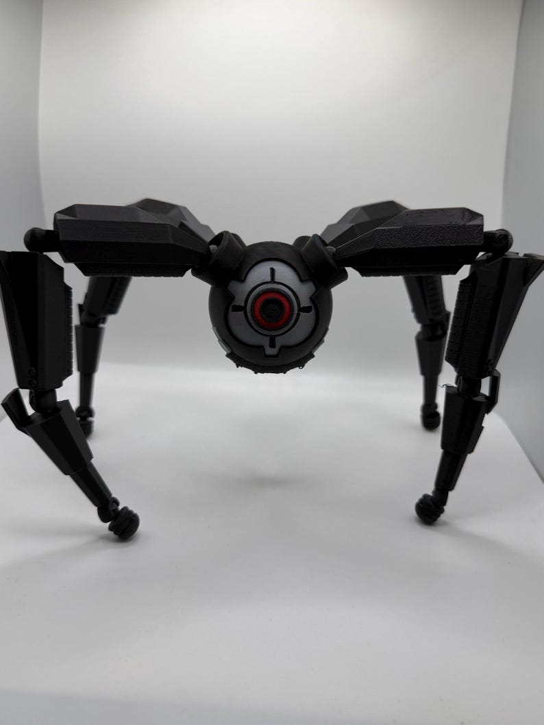 May include: A black, mechanical spider-like robot with a central eye-like sensor. The robot has multiple articulated legs and a detailed, industrial design. The eye-like sensor is gray with a red center.