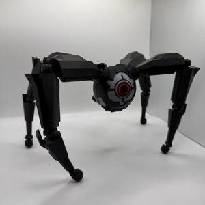 May include: A black, mechanical spider toy with articulated legs and a central body featuring a circular eye with red and white details. The toy is set against a plain white background.