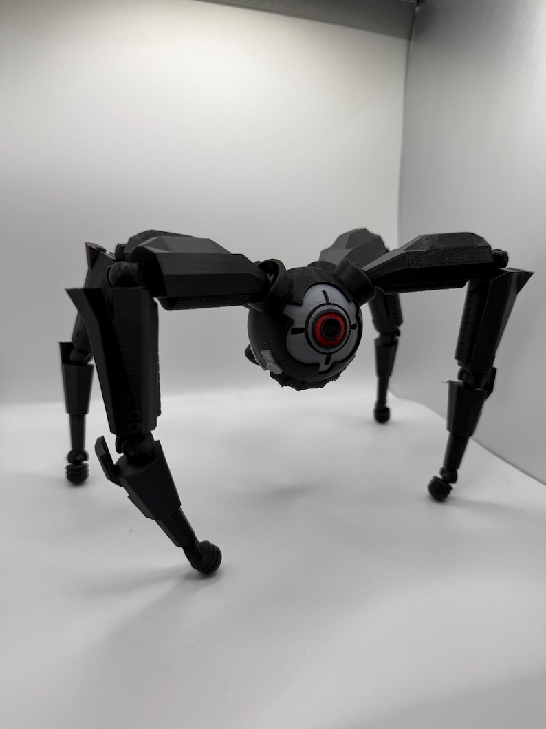 May include: A black, mechanical spider toy with articulated legs and a central body featuring a circular eye with red and white details. The toy is set against a plain white background.