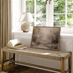 May include: A light brown wooden bench with a woven seat and a book on top. The bench is in front of a window with a white frame and green trees outside. A lamp with a white shade and a brown ceramic base is on the windowsill. A framed painting with a brown background is on the wall above the bench.