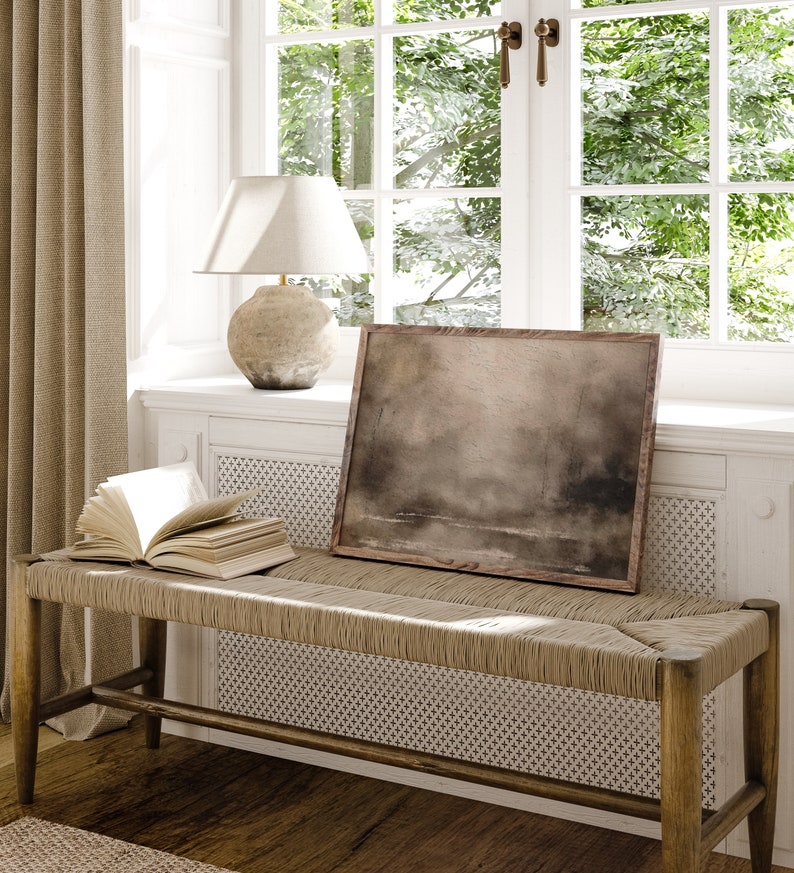 May include: A light brown wooden bench with a woven seat and a book on top. The bench is in front of a window with a white frame and green trees outside. A lamp with a white shade and a brown ceramic base is on the windowsill. A framed painting with a brown background is on the wall above the bench.