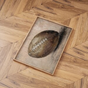 May include: A framed watercolor painting of a brown leather football on a light brown background.