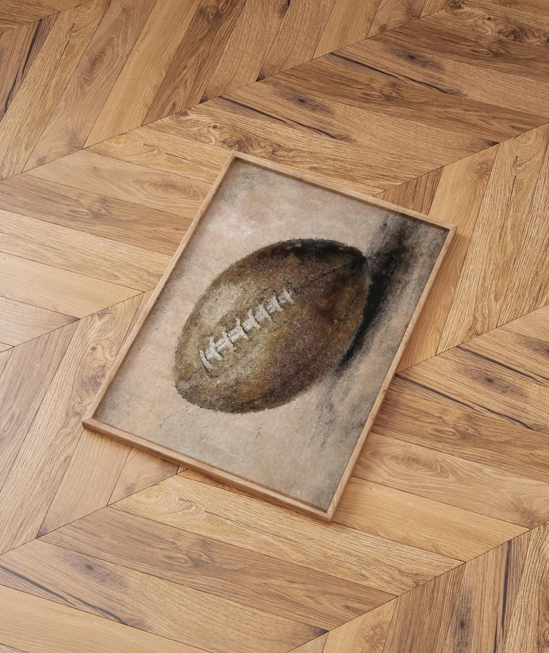 May include: A framed watercolor painting of a brown leather football on a light brown background.