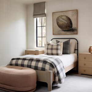 May include: A bedroom with a white and black plaid blanket on a bed with a black metal frame. There is a brown football painting on the wall above the bed. There is a light pink ottoman in the foreground.