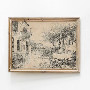 Puede incluir: Un boceto a lápiz enmarcado de una escena de café europeo. La obra de arte representa un edificio con balcón, un árbol, mesas y sillas. El marco es de color marrón claro.