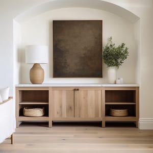 May include: A light wood cabinet with open shelving and closed doors, topped with a marble surface. A large framed abstract art piece hangs above the cabinet. A lamp with a beige base and white shade sits on the cabinet.