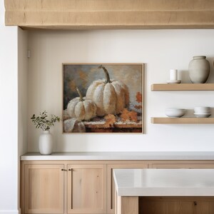 Pode incluir: Uma pintura emoldurada de duas ab&oacute;boras brancas com folhas laranja sobre uma toalha de mesa branca. A pintura est&aacute; pendurada em uma parede branca acima de uma bancada de cozinha com arm&aacute;rios de madeira.