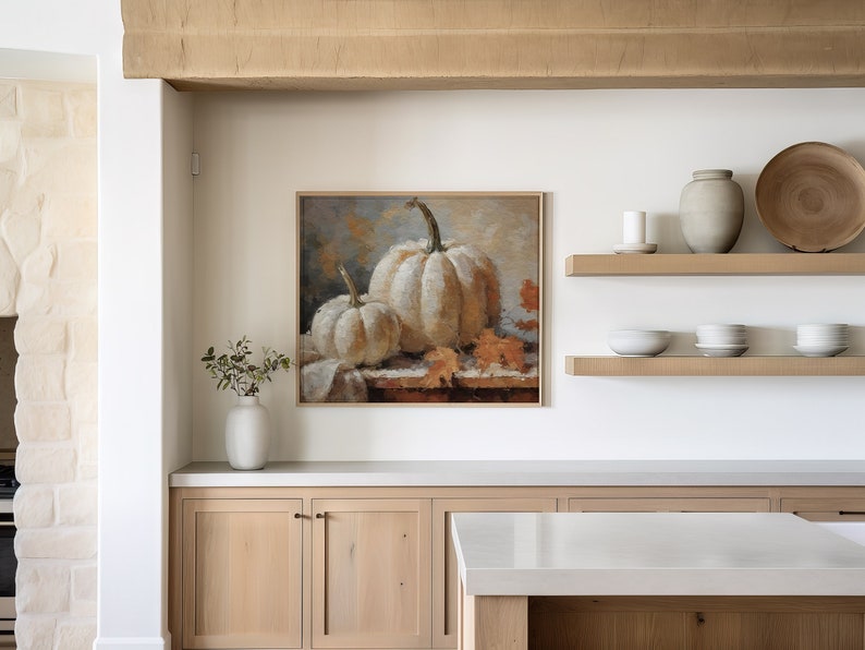 Pode incluir: Uma pintura emoldurada de duas ab&oacute;boras brancas com folhas laranja sobre uma toalha de mesa branca. A pintura est&aacute; pendurada em uma parede branca acima de uma bancada de cozinha com arm&aacute;rios de madeira.