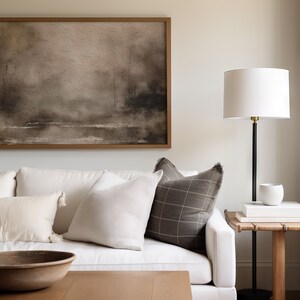 May include: A white couch with a brown and white plaid pillow, a white pillow, and a wooden bowl on a coffee table in front of it. A framed abstract painting hangs on the wall behind the couch. A white lamp with a black stand sits on a wooden side table next to the couch.