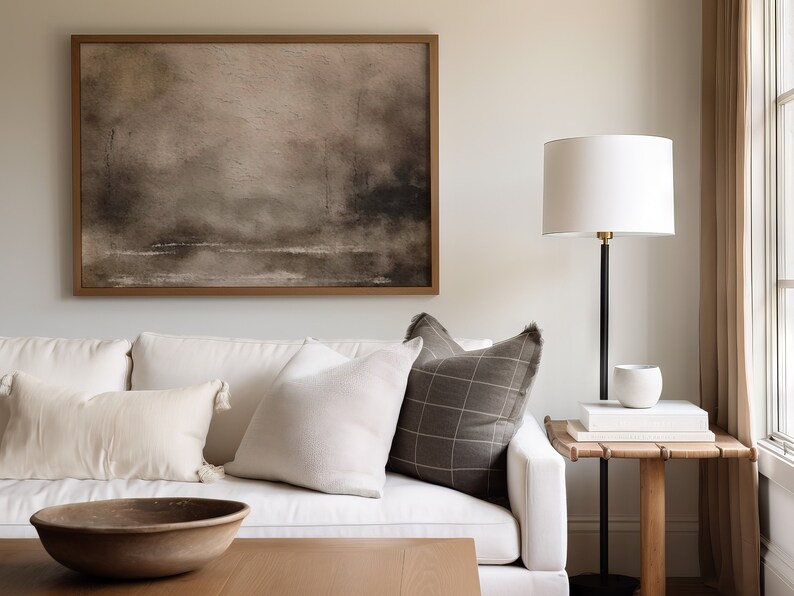 May include: A white couch with a brown and white plaid pillow, a white pillow, and a wooden bowl on a coffee table in front of it. A framed abstract painting hangs on the wall behind the couch. A white lamp with a black stand sits on a wooden side table next to the couch.