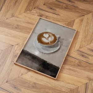 K&ouml;nnte beinhalten: Ein gerahmtes Gem&auml;lde einer Tasse Kaffee mit Latte-Art-Design. Der Kaffee befindet sich in einer wei&szlig;en Tasse und Untertasse auf einer hellgrauen Oberfl&auml;che.