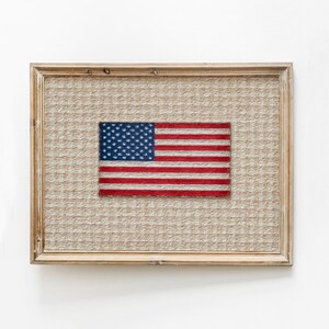 May include: Framed artwork featuring a textured background and an embroidered American flag. The flag displays a blue field with white stars and red and white stripes. The frame is made of light-colored wood.