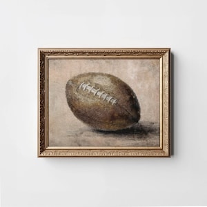 May include: A vintage-style painting of a brown leather football in a gold frame. The football is slightly worn and has white stitching.