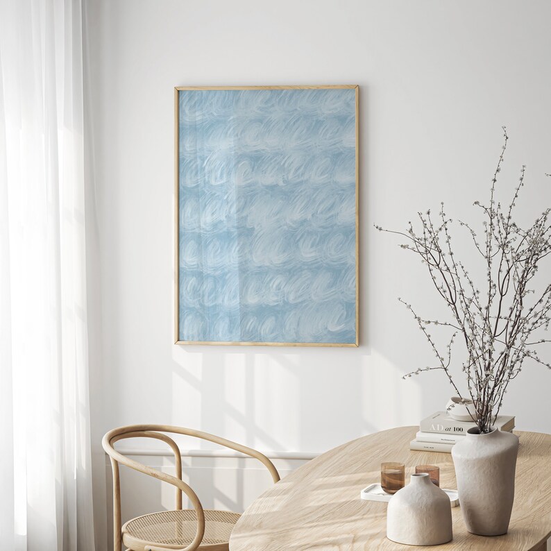 May include: A light blue abstract painting in a gold frame hangs on a white wall above a wooden table with a vase of white branches and a book.