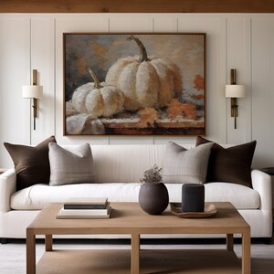 Pode incluir: Um sof&aacute; branco com almofadas marrons fica em uma sala de estar com uma mesa de centro e uma pintura de ab&oacute;boras brancas na parede.