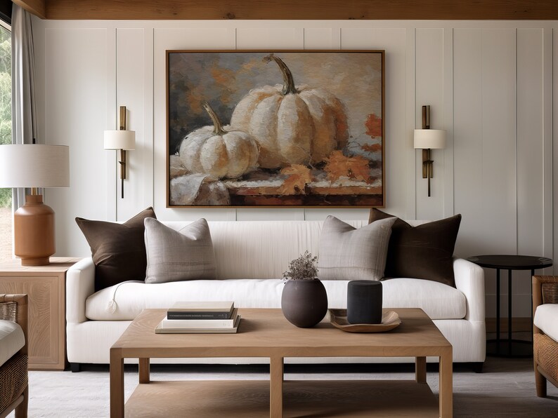Pode incluir: Um sof&aacute; branco com almofadas marrons fica em uma sala de estar com uma mesa de centro e uma pintura de ab&oacute;boras brancas na parede.