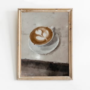 K&ouml;nnte beinhalten: Ein gerahmtes Gem&auml;lde einer Tasse Kaffee mit Latte Art in Form eines Blattes. Der Kaffee befindet sich in einer wei&szlig;en Tasse und Untertasse auf einer hellbraunen Oberfl&auml;che.