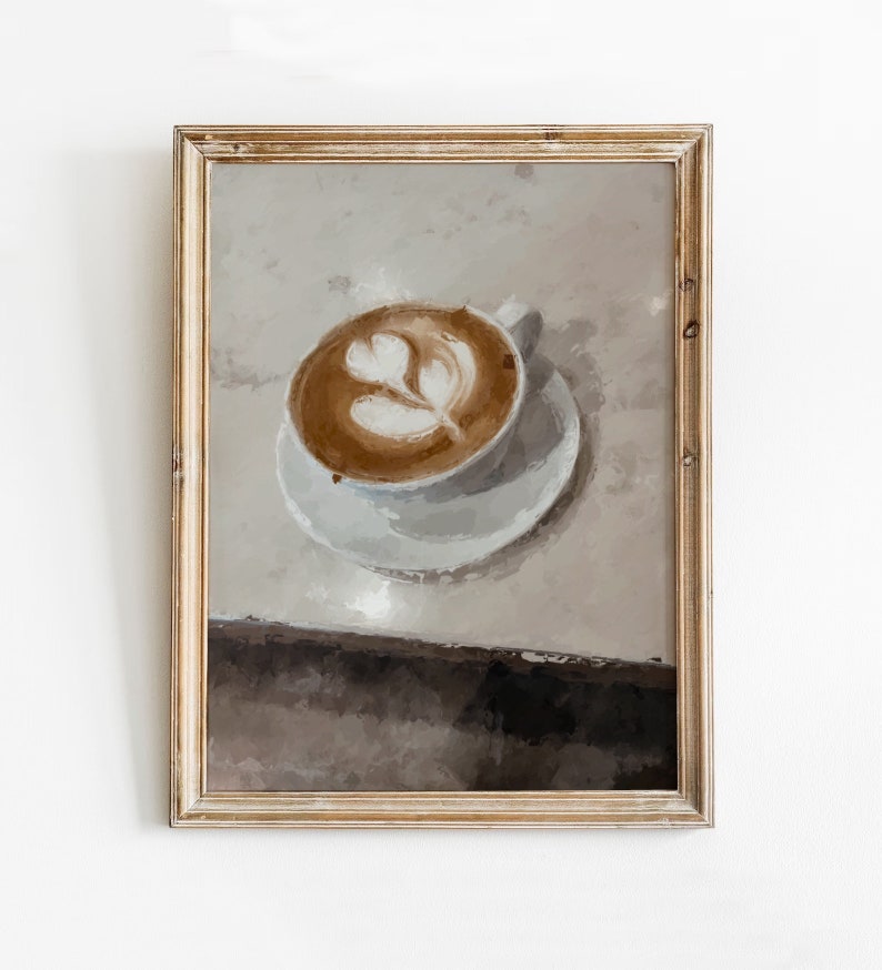 K&ouml;nnte beinhalten: Ein gerahmtes Gem&auml;lde einer Tasse Kaffee mit Latte Art in Form eines Blattes. Der Kaffee befindet sich in einer wei&szlig;en Tasse und Untertasse auf einer hellbraunen Oberfl&auml;che.