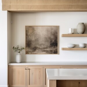 May include: A white kitchen with light wood cabinets and a white countertop. There is a framed abstract painting on the wall above the cabinets. Two wooden shelves with decorative items are on the wall to the right of the painting.