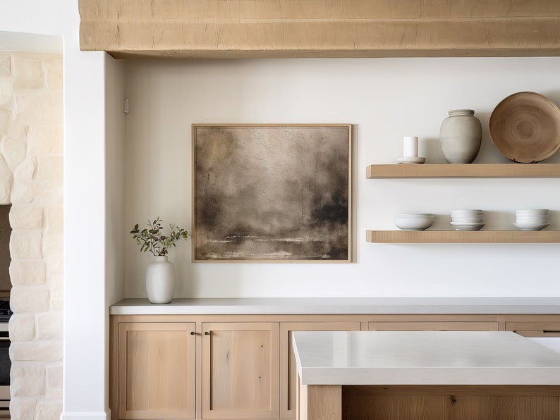 May include: A white kitchen with light wood cabinets and a white countertop. There is a framed abstract painting on the wall above the cabinets. Two wooden shelves with decorative items are on the wall to the right of the painting.