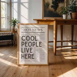 Puede incluir: Un cartel enmarcado con el titular "HOT NEWS" y el texto "COOL PEOPLE LIVE HERE" en un dise&ntilde;o de estilo peri&oacute;dico. El cartel est&aacute; en un marco de madera clara, apoyado contra un suelo de madera. La habitaci&oacute;n tiene muebles de madera y plantas en macetas.