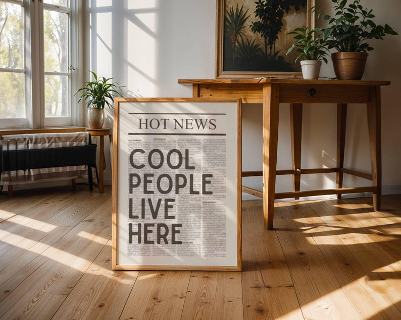 Puede incluir: Un cartel enmarcado con el titular "HOT NEWS" y el texto "COOL PEOPLE LIVE HERE" en un dise&ntilde;o de estilo peri&oacute;dico. El cartel est&aacute; en un marco de madera clara, apoyado contra un suelo de madera. La habitaci&oacute;n tiene muebles de madera y plantas en macetas.