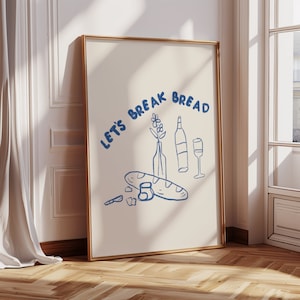 May include: A beige poster with a gold frame featuring a blue illustration of a baguette, wine bottle, wine glass, and flowers with the text "Let's Break Bread".