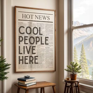 Puede incluir: Un marco de madera que contiene una impresi&oacute;n de peri&oacute;dico con el titular "HOT NEWS" y el texto "COOL PEOPLE LIVE HERE". Una peque&ntilde;a mesa de madera con libros y una c&aacute;mara se encuentra debajo, cerca de una ventana.