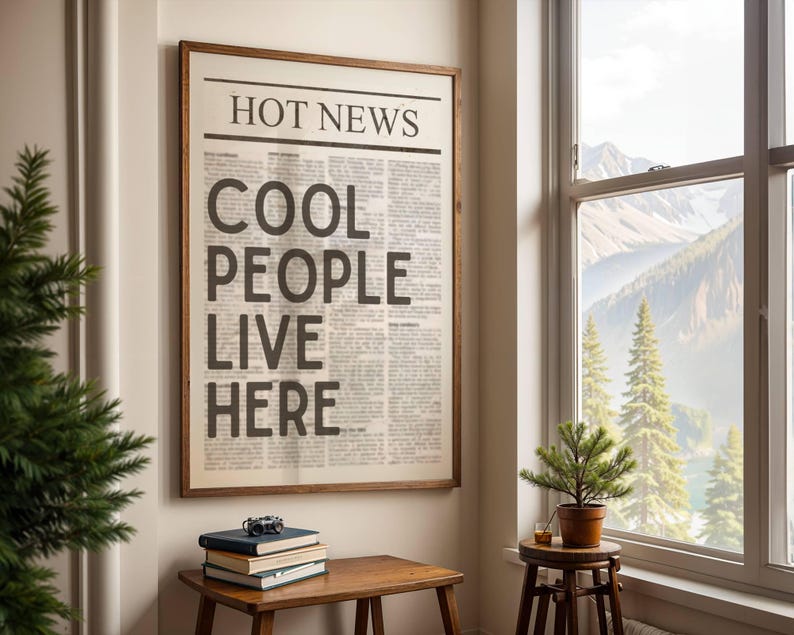 Puede incluir: Un marco de madera que contiene una impresi&oacute;n de peri&oacute;dico con el titular "HOT NEWS" y el texto "COOL PEOPLE LIVE HERE". Una peque&ntilde;a mesa de madera con libros y una c&aacute;mara se encuentra debajo, cerca de una ventana.
