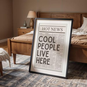 Puede incluir: Un cartel enmarcado con el texto "HOT NEWS COOL PEOPLE LIVE HERE" en estilo peri&oacute;dico. El marco negro se apoya en una alfombra estampada en un dormitorio, con una cama de madera y una mesita de noche visibles.