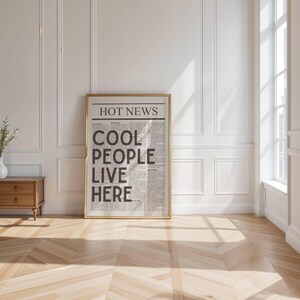 Puede incluir: Un cartel enmarcado con el texto "HOT NEWS COOL PEOPLE LIVE HERE" en un dise&ntilde;o de peri&oacute;dico. El cartel est&aacute; en un marco dorado y se apoya en una pared blanca. Un aparador de madera con un jarr&oacute;n blanco y vegetaci&oacute;n est&aacute; a la izquierda.