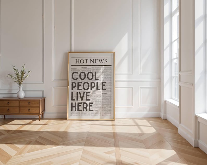 Puede incluir: Un cartel enmarcado con el texto "HOT NEWS COOL PEOPLE LIVE HERE" en un dise&ntilde;o de peri&oacute;dico. El cartel est&aacute; en un marco dorado y se apoya en una pared blanca. Un aparador de madera con un jarr&oacute;n blanco y vegetaci&oacute;n est&aacute; a la izquierda.