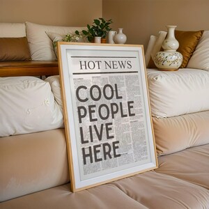 Puede incluir: Un marco con el titular "HOT NEWS" y el texto "COOL PEOPLE LIVE HERE" en negrita, letras negras. La impresi&oacute;n est&aacute; en un marco de madera clara y descansa contra un sof&aacute; beige con cojines blancos y jarrones decorativos.