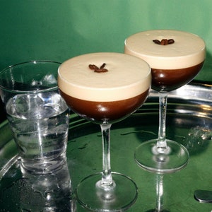 May include: Two clear glass martini glasses filled with a layered coffee drink. The bottom layer is dark brown coffee, and the top layer is a light brown, foamy cream. Each glass has a few coffee beans on top of the cream. The glasses are on a silver tray with a green background.