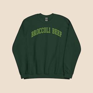 May include: A dark green sweatshirt featuring the text "BROCCOLI BEEF" in a light green, curved font. The long-sleeved sweatshirt has a crew neck. The garment is a solid colour and appears to be made of a soft material.