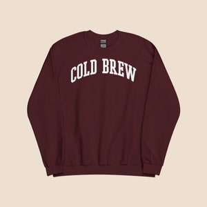 Op de afbeelding: Een bordeauxrood sweatshirt met de tekst "COLD BREW" in wit, in gebogen letters.